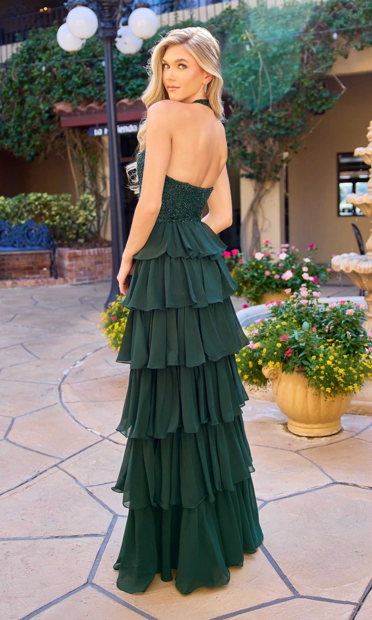 A woman in the Primavera 4530 Long Prom Dress, a strapless dark green gown with a tiered ruffle chiffon skirt, stands outdoors on a stone path surrounded by plants, flowers, and a lamppost, looking over her shoulder.